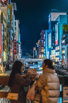 A vibrant urban night scene featuring a bustling canal lined with illuminated buildings displaying colorful signs. Two people in the foreground appear to be in conversation, adding a sense of lively interaction to the setting.