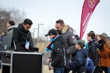 A group of people, including children and adults, are gathered outdoors near a kiosk. One person appears to be an event staff member checking tickets or passes. The attendees are dressed warmly, indicating cooler weather. In the background, a red flag with a logo and the words 'Official Programme' is visible, suggesting an organized event or sports game atmosphere.