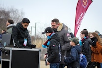 A group of people, including children and adults, are gathered outdoors near a kiosk. One person appears to be an event staff member checking tickets or passes. The attendees are dressed warmly, indicating cooler weather. In the background, a red flag with a logo and the words 'Official Programme' is visible, suggesting an organized event or sports game atmosphere.