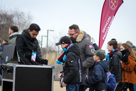 A group of people, including children and adults, are gathered outdoors near a kiosk. One person appears to be an event staff member checking tickets or passes. The attendees are dressed warmly, indicating cooler weather. In the background, a red flag with a logo and the words 'Official Programme' is visible, suggesting an organized event or sports game atmosphere.