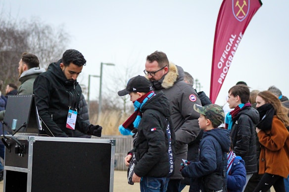A group of people, including children and adults, are gathered outdoors near a kiosk. One person appears to be an event staff member checking tickets or passes. The attendees are dressed warmly, indicating cooler weather. In the background, a red flag with a logo and the words 'Official Programme' is visible, suggesting an organized event or sports game atmosphere.