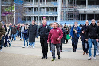 A bustling crowd of people walking outside, with some wearing sports jerseys. The background features an urban environment with construction scaffoldings. The atmosphere is lively and casual, suggesting perhaps an event or gathering.