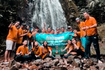 A group of people wearing similar orange shirts are gathered in front of a waterfall, holding a large banner. The scene suggests a team outing or group event in a natural setting with rocks and greenery around.