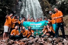 A group of people wearing similar orange shirts are gathered in front of a waterfall, holding a large banner. The scene suggests a team outing or group event in a natural setting with rocks and greenery around.