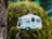 A birdhouse designed to look like a vintage camper van with detailed features including windows, a door, and a spare tire. It hangs from a rope against a backdrop of blurred greenery, providing a whimsical and nostalgic aesthetic.