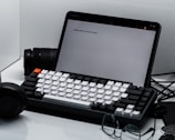 A neatly arranged workspace featuring a tablet with text displayed on the screen, alongside a mechanical keyboard with white and black keys. In the background, a camera lens is partially visible. A pair of black headphones and a pair of eyeglasses are placed nearby on the clean, white surface.