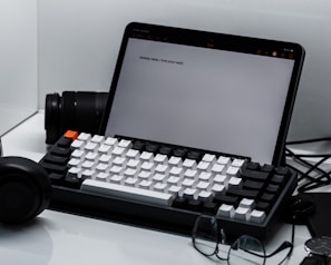 A neatly arranged workspace featuring a tablet with text displayed on the screen, alongside a mechanical keyboard with white and black keys. In the background, a camera lens is partially visible. A pair of black headphones and a pair of eyeglasses are placed nearby on the clean, white surface.