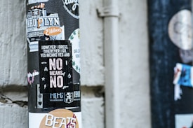 A collection of layered stickers on a cylindrical pole against a concrete wall. The most prominent sticker reads 'However I dress, wherever I go, Yes means yes and No means no' in bold letters, surrounded by various graphics and logos. Other stickers display text and images related to music and sports.