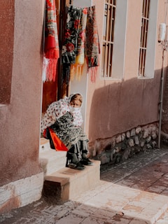An elderly person wearing traditional clothing sits on a step outside a building. The individual is wrapped in a white patterned blanket and is positioned under vibrant embroidered textiles hanging on the wall. The scene is bright with natural sunlight casting shadows on the pavement.