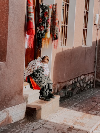 An elderly person wearing traditional clothing sits on a step outside a building. The individual is wrapped in a white patterned blanket and is positioned under vibrant embroidered textiles hanging on the wall. The scene is bright with natural sunlight casting shadows on the pavement.