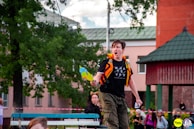 A person with a determined expression is holding a water gun and appears to be shouting while wearing a black t-shirt with the word 'WHATEVER' printed on it. The scene is outdoors with several other people in the background, some dressed in costumes, near a tree and a building with a red and green roof.