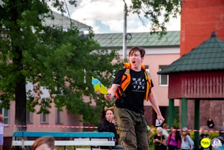 A person with a determined expression is holding a water gun and appears to be shouting while wearing a black t-shirt with the word 'WHATEVER' printed on it. The scene is outdoors with several other people in the background, some dressed in costumes, near a tree and a building with a red and green roof.