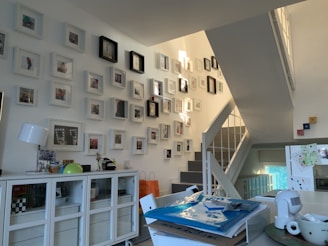 A modern, well-lit interior space featuring a collection of framed photos mounted on a staircase wall. The room includes a white sideboard with decorative items such as a lamp and globe. Several stacked papers and a cup are placed on a table, suggesting a lived-in, organized environment.