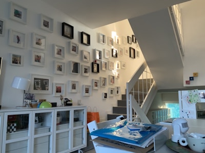 A modern, well-lit interior space featuring a collection of framed photos mounted on a staircase wall. The room includes a white sideboard with decorative items such as a lamp and globe. Several stacked papers and a cup are placed on a table, suggesting a lived-in, organized environment.