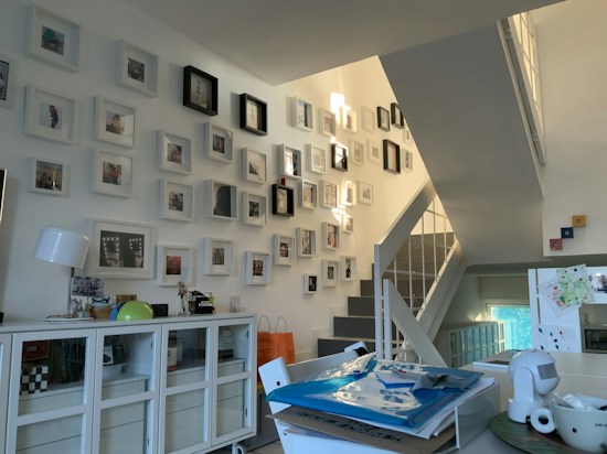 A modern, well-lit interior space featuring a collection of framed photos mounted on a staircase wall. The room includes a white sideboard with decorative items such as a lamp and globe. Several stacked papers and a cup are placed on a table, suggesting a lived-in, organized environment.
