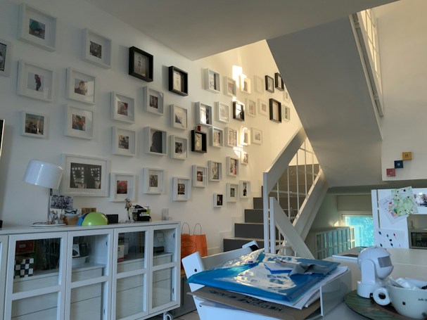 A modern, well-lit interior space featuring a collection of framed photos mounted on a staircase wall. The room includes a white sideboard with decorative items such as a lamp and globe. Several stacked papers and a cup are placed on a table, suggesting a lived-in, organized environment.