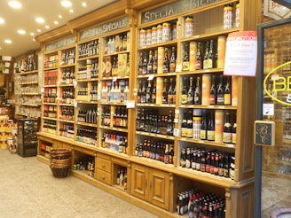 A well-stocked beer store shelf featuring a wide variety of beer bottles and cans organized in sections. The wooden shelves have a classic look, decorated with chalkboard signs indicating different beer categories. Promotional signs are displayed, and a basket on the floor holds additional items.