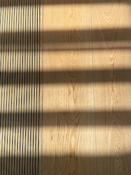 Wooden flooring with alternating vertical slats on the left side. Shadows cast across the surface, creating a pattern of light and dark bands.