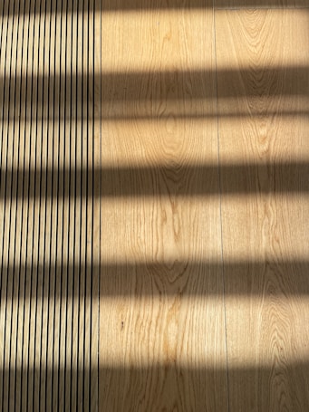 Wooden flooring with alternating vertical slats on the left side. Shadows cast across the surface, creating a pattern of light and dark bands.