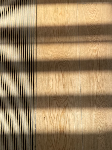 Wooden flooring with alternating vertical slats on the left side. Shadows cast across the surface, creating a pattern of light and dark bands.