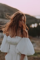 A woman with long, flowing hair wears a white off-the-shoulder dress with voluminous sleeves. The background features a soft, blurred landscape, possibly during sunset, creating a serene atmosphere.