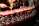 A bartender is pouring a reddish cocktail through a strainer into a row of elegant glasses lined up on a bar counter.