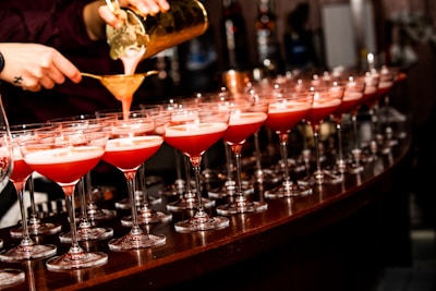 A bartender is pouring a reddish cocktail through a strainer into a row of elegant glasses lined up on a bar counter.
