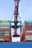 A large industrial crane is positioned over a stack of colorful shipping containers, lifting one container. The containers are branded with various shipping company names and appear to be part of a busy port operation.