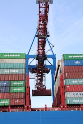 A large industrial crane is positioned over a stack of colorful shipping containers, lifting one container. The containers are branded with various shipping company names and appear to be part of a busy port operation.