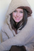 A person dressed in a warm, fluffy coat with a hood, which is beige with a brown interior lining. The individual has long, dark hair and is smiling, suggesting a relaxed and comfortable demeanor.