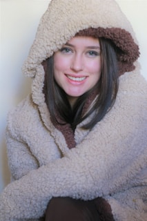 A person dressed in a warm, fluffy coat with a hood, which is beige with a brown interior lining. The individual has long, dark hair and is smiling, suggesting a relaxed and comfortable demeanor.