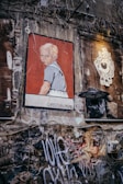 A street art mural on a weathered wall depicts a young boy with blond hair in a white shirt and overalls, set against a red background. The name 'Christian Paine' is inscribed below the image. Nearby, a stylized eye graphic is visible next to some exposed wires, and the wall is covered with various graffiti tags and textures.