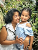 A woman and a child are smiling together in an outdoor setting with lush green foliage in the background. The woman is wearing a white polka dot dress, and the child is dressed in a light blue dress with white lace details. They appear to be enjoying a moment of connection and happiness.