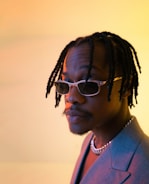 A person with braided hair and sunglasses gazes thoughtfully off-camera. They are wearing a gray blazer and a chain necklace, set against a softly lit background with warm yellow and orange hues.