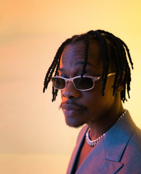 A person with braided hair and sunglasses gazes thoughtfully off-camera. They are wearing a gray blazer and a chain necklace, set against a softly lit background with warm yellow and orange hues.