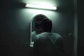 A person facing a wall-mounted mirror is illuminated by an overhead fluorescent light. The room appears to have dim lighting, creating a moody atmosphere.