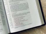 An open Bible is displayed on a wooden surface, revealing text from the New Testament. The left page includes sections on Jesus healing a great multitude, while the right page highlights the Beatitudes from the Sermon on the Mount, with text in red and black for emphasis. The text is well-arranged in columns with chapter and verse references.