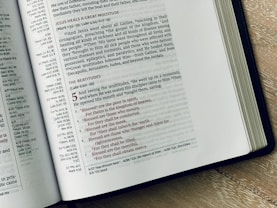 An open Bible is displayed on a wooden surface, revealing text from the New Testament. The left page includes sections on Jesus healing a great multitude, while the right page highlights the Beatitudes from the Sermon on the Mount, with text in red and black for emphasis. The text is well-arranged in columns with chapter and verse references.