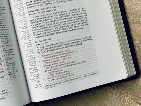An open Bible is displayed on a wooden surface, revealing text from the New Testament. The left page includes sections on Jesus healing a great multitude, while the right page highlights the Beatitudes from the Sermon on the Mount, with text in red and black for emphasis. The text is well-arranged in columns with chapter and verse references.
