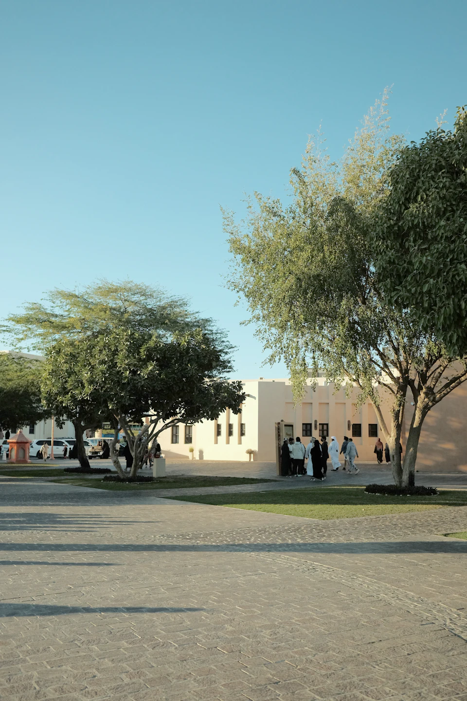 The image depicts a serene courtyard scene with a large white building featuring arched windows and a flat roof, surrounded by lush green trees. The courtyard is paved with gray cobblestones and features several people walking around or standing near the buildings.