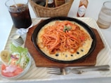 A neatly arranged meal placed on a rectangular tray, featuring spaghetti topped with green peas on a sizzling plate with a cheesy base. Accompanying the main dish is a side salad with lettuce, tomato slices, a slice of banana, and a creamy dressing served in a small jug. A glass of iced cola, a bottle of Tabasco sauce, and a basket containing condiments are also present.