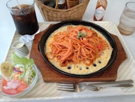 A neatly arranged meal placed on a rectangular tray, featuring spaghetti topped with green peas on a sizzling plate with a cheesy base. Accompanying the main dish is a side salad with lettuce, tomato slices, a slice of banana, and a creamy dressing served in a small jug. A glass of iced cola, a bottle of Tabasco sauce, and a basket containing condiments are also present.