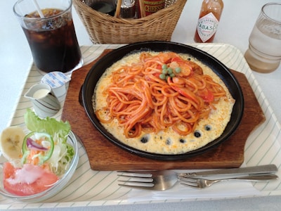A neatly arranged meal placed on a rectangular tray, featuring spaghetti topped with green peas on a sizzling plate with a cheesy base. Accompanying the main dish is a side salad with lettuce, tomato slices, a slice of banana, and a creamy dressing served in a small jug. A glass of iced cola, a bottle of Tabasco sauce, and a basket containing condiments are also present.