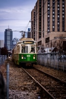 A green trolley with an accessibility symbol approaches on a section of urban light rail track, flanked by chain-link fences. In the background, tall city buildings are visible under a cloudy sky, with additional modern architecture further enhancing the urban setting.