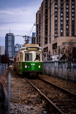 A green trolley with an accessibility symbol approaches on a section of urban light rail track, flanked by chain-link fences. In the background, tall city buildings are visible under a cloudy sky, with additional modern architecture further enhancing the urban setting.