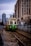 A green trolley with an accessibility symbol approaches on a section of urban light rail track, flanked by chain-link fences. In the background, tall city buildings are visible under a cloudy sky, with additional modern architecture further enhancing the urban setting.