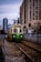 A green trolley with an accessibility symbol approaches on a section of urban light rail track, flanked by chain-link fences. In the background, tall city buildings are visible under a cloudy sky, with additional modern architecture further enhancing the urban setting.