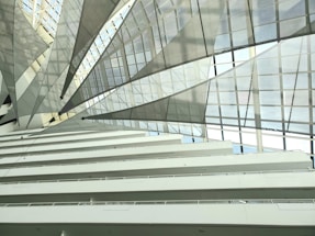 Interior view of a modern building with multiple floors and a large, geometric, glass-paneled roof with an intricate architectural design. The roof features translucent panels and metallic supports, creating a dynamic, airy feel.