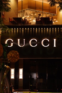 A dimly lit shop front displaying the illuminated Gucci logo in large letters. Above the sign, a warm-lit interior with tables and chairs is visible through the glass, suggesting a stylish setting. Decorative elements and fixtures enhance the luxurious atmosphere.