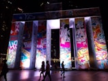 A brightly lit art installation with large panels covered in colorful, abstract designs and graphic imagery. Silhouettes of people walking in the foreground contrast with the vibrant lights.